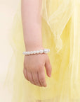 A close-up of a child's hand and wrist wearing a beaded bracelet made of white faux pearls, styled with a soft yellow tulle dress against a light cream background.