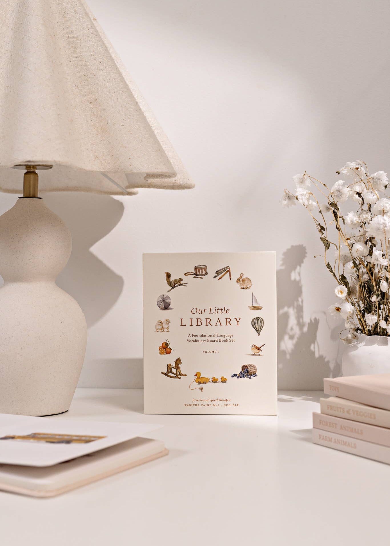 Book titled 'Our Little Library' on a table with a lamp and flowers in the background