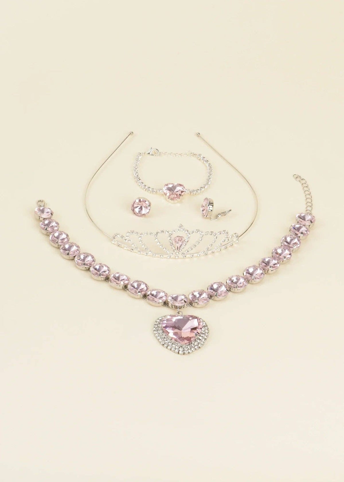 A pink and silver jewelry set on a light cream background, featuring a rhinestone-embellished collar necklace with a heart-shaped pendant, a tiara, a bracelet, and matching stud earrings.