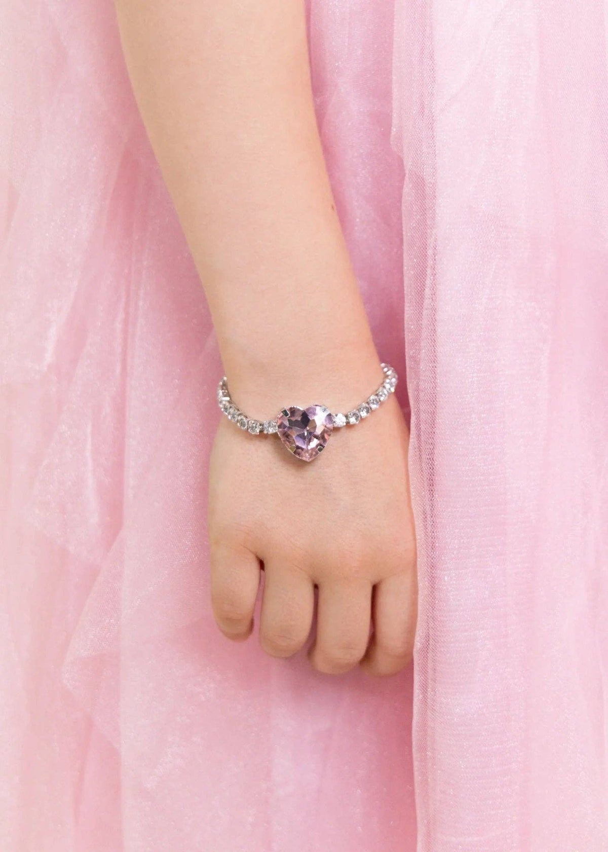 A close-up of a child's hand and wrist wearing a delicate silver-toned bracelet with a heart-shaped purple gemstone, set against a soft pink tulle dress background.