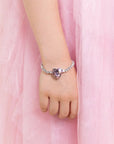 A close-up of a child's hand and wrist wearing a delicate silver-toned bracelet with a heart-shaped purple gemstone, set against a soft pink tulle dress background.