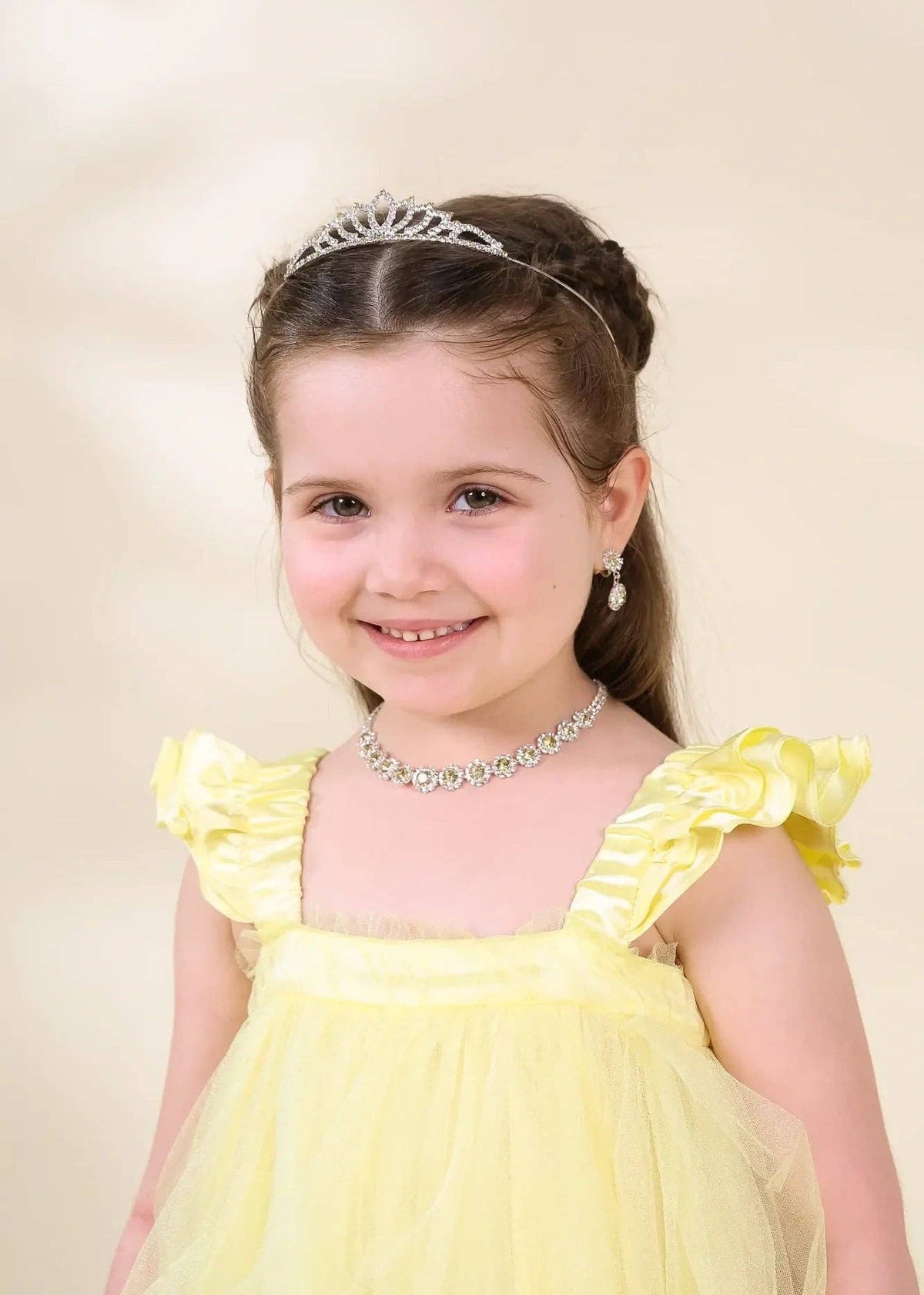 A young girl with brown hair in a half-up style wearing a silver tiara, matching silver necklace, and a yellow dress with ruffled shoulders, photographed against a soft beige studio background.