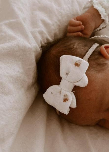 Baby wearing a white headband with a bow, lying on a soft beige blanket.