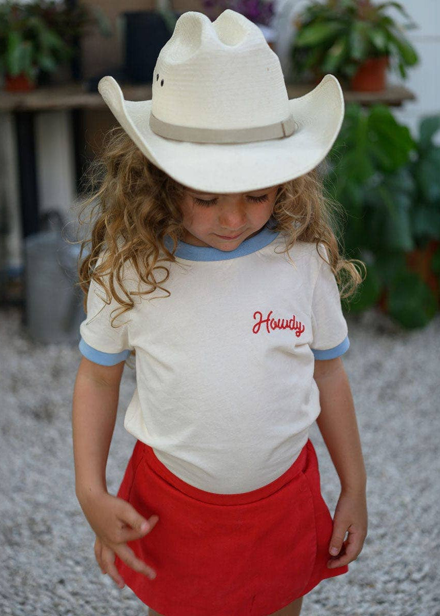 Child wearing a cowboy hat and 'Howdy' shirt with plants in the background