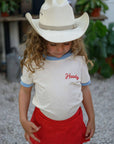 Child wearing a cowboy hat and 'Howdy' shirt with plants in the background