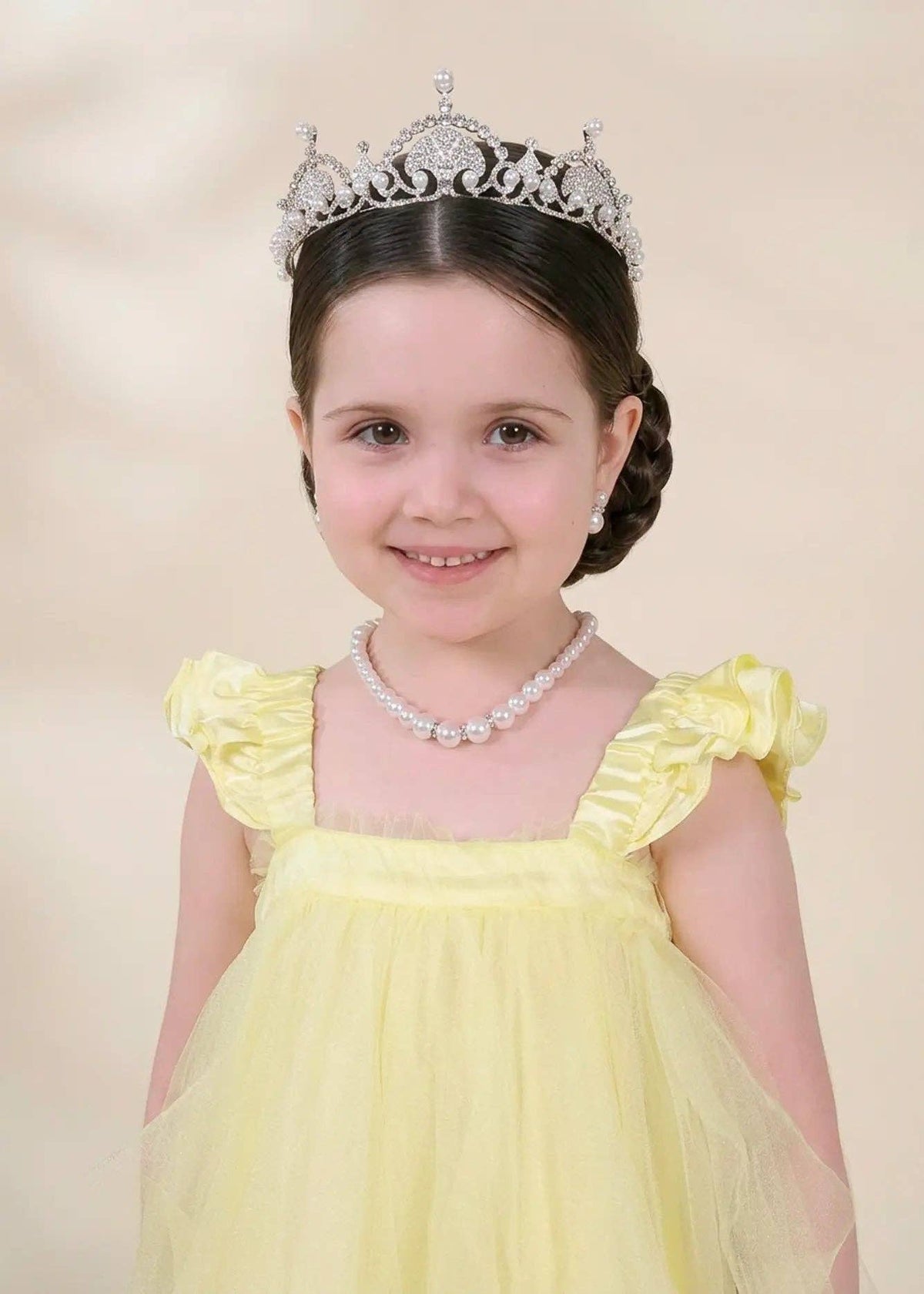 A young girl with dark hair styled in a braid, wearing a silver tiara, a pearl necklace, and a pale yellow dress with ruffled shoulders, set against a soft beige background.
