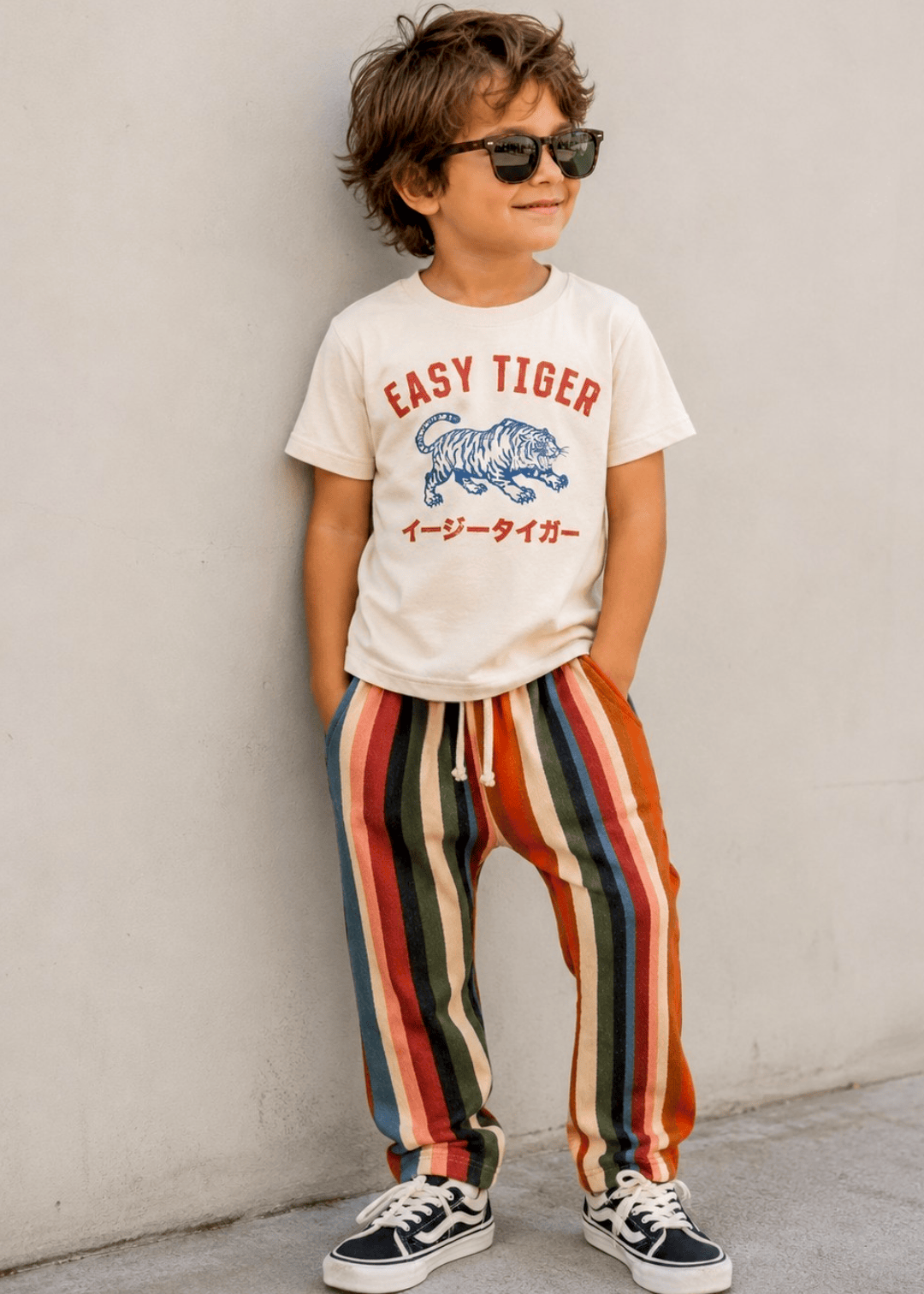 Child wearing a 'Easy Tiger' t-shirt and colorful striped pants against a plain wall.