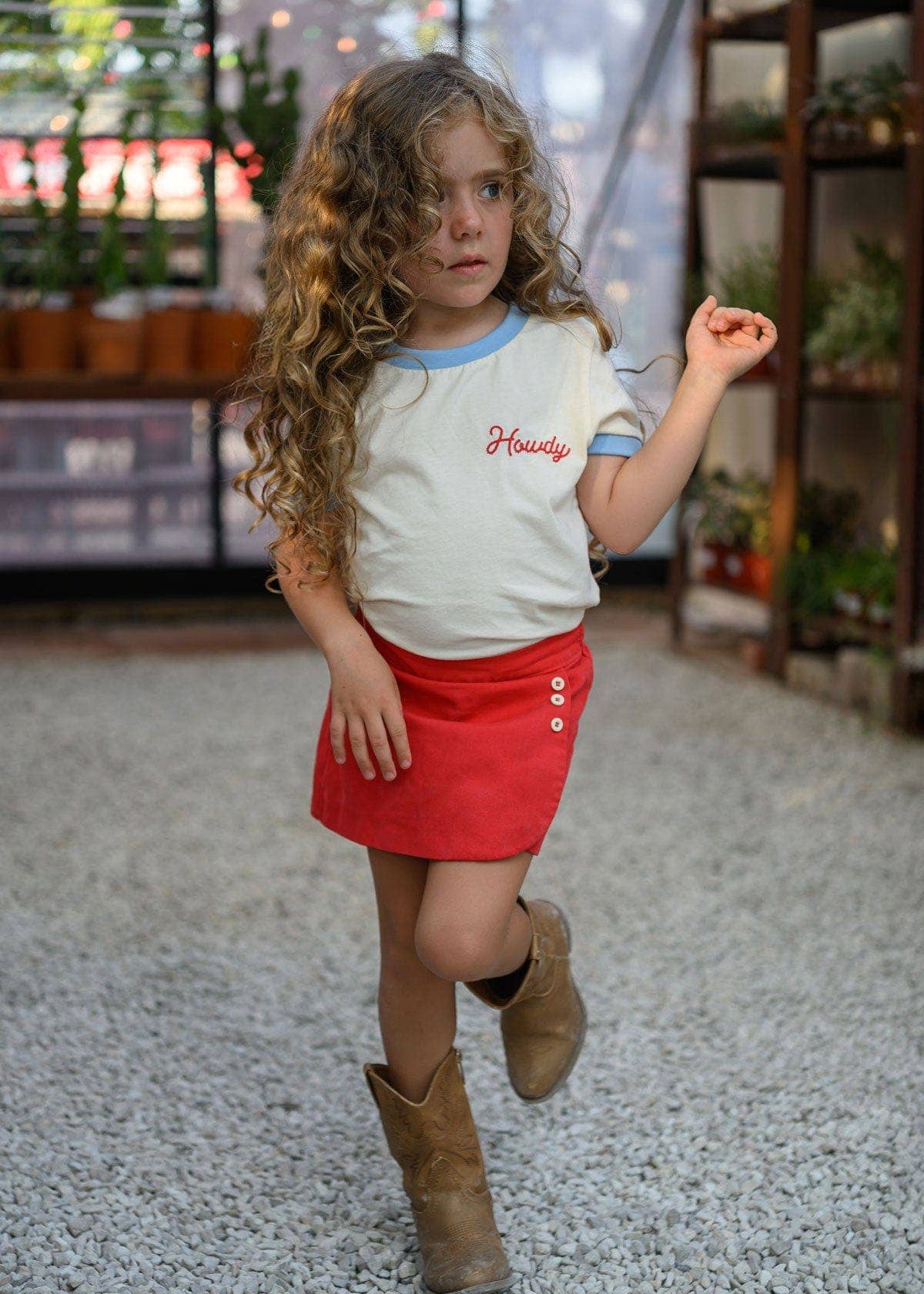 Child wearing a white shirt with text and red skirt, standing on a gravel path.