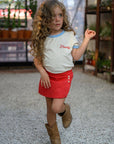 Child wearing a white shirt with text and red skirt, standing on a gravel path.