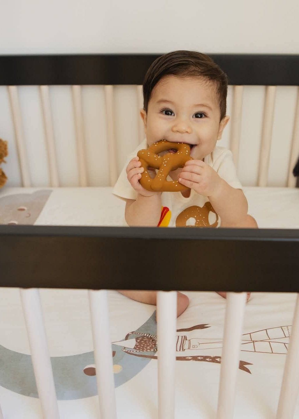 A baby sitting in a black and white crib, holding a brown plush toy. The crib is lined with a white patterned sheet and surrounded by various stuffed animals, including a panda and a bear.