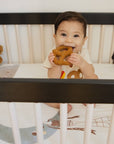 A baby sitting in a black and white crib, holding a brown plush toy. The crib is lined with a white patterned sheet and surrounded by various stuffed animals, including a panda and a bear.
