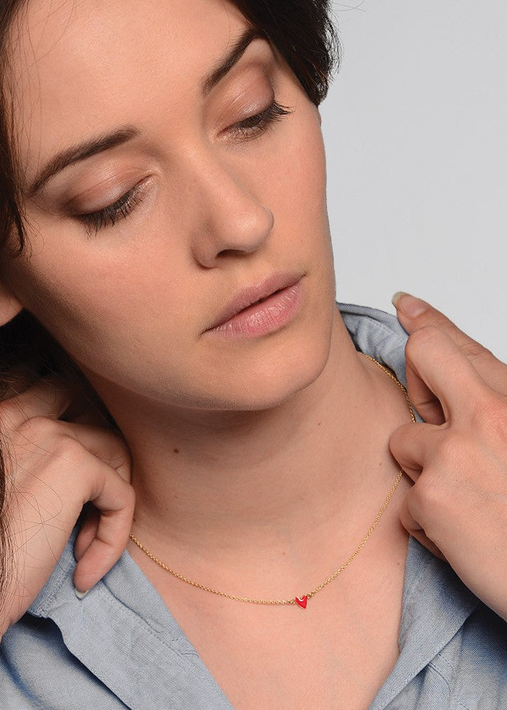 Woman wearing a gold necklace with a red heart pendant, with hands gently touching her shoulders.