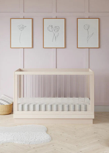 Wooden crib in a room with light pink walls, framed artwork, and decorative items.