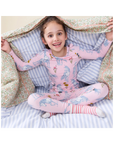 A young girl smiling while sitting cross-legged under a patterned blanket, wearing a pink long-sleeve pajama set with a blue unicorn print and striped socks.