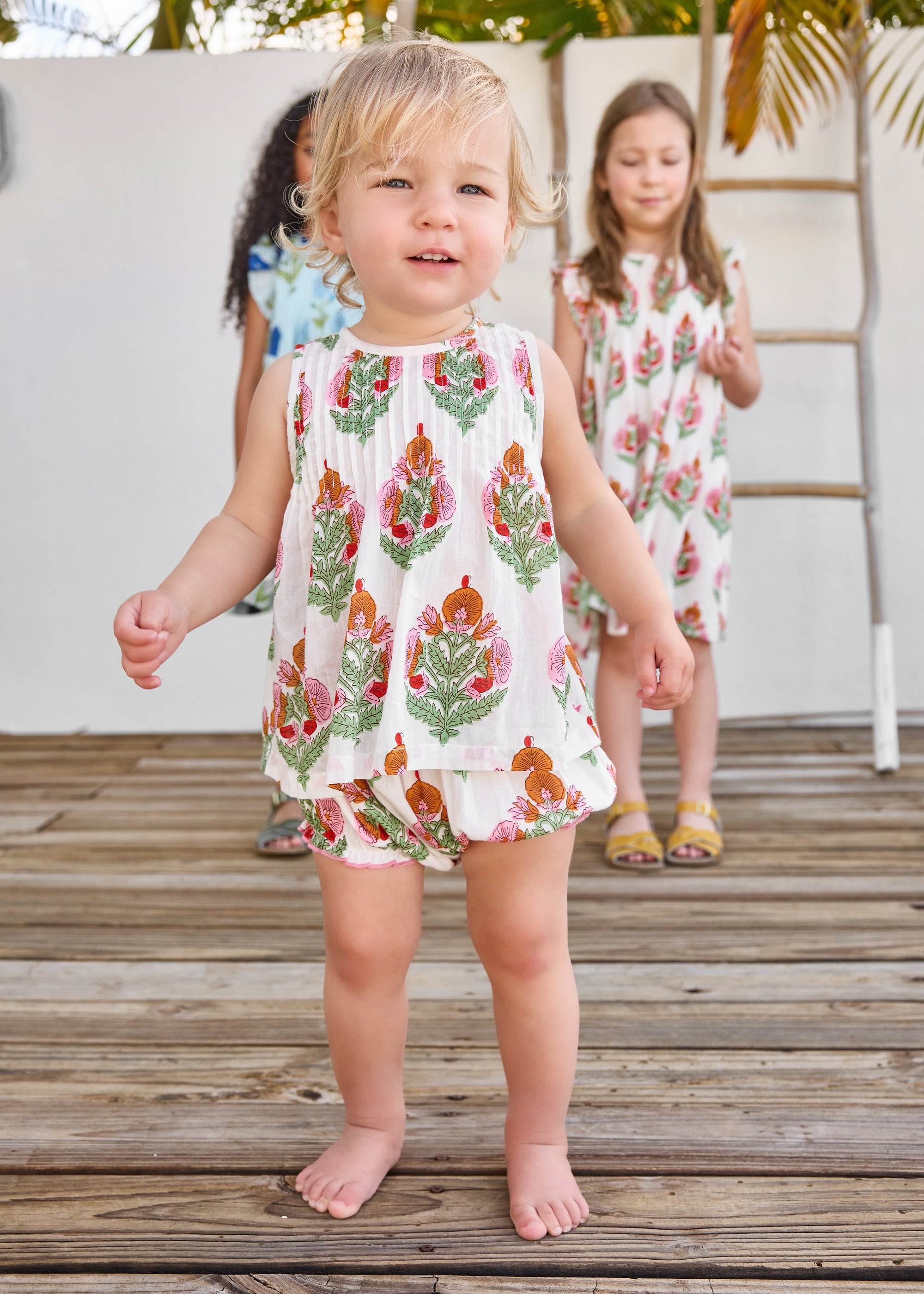 Two young girls in floral dresses standing on a wooden deck.