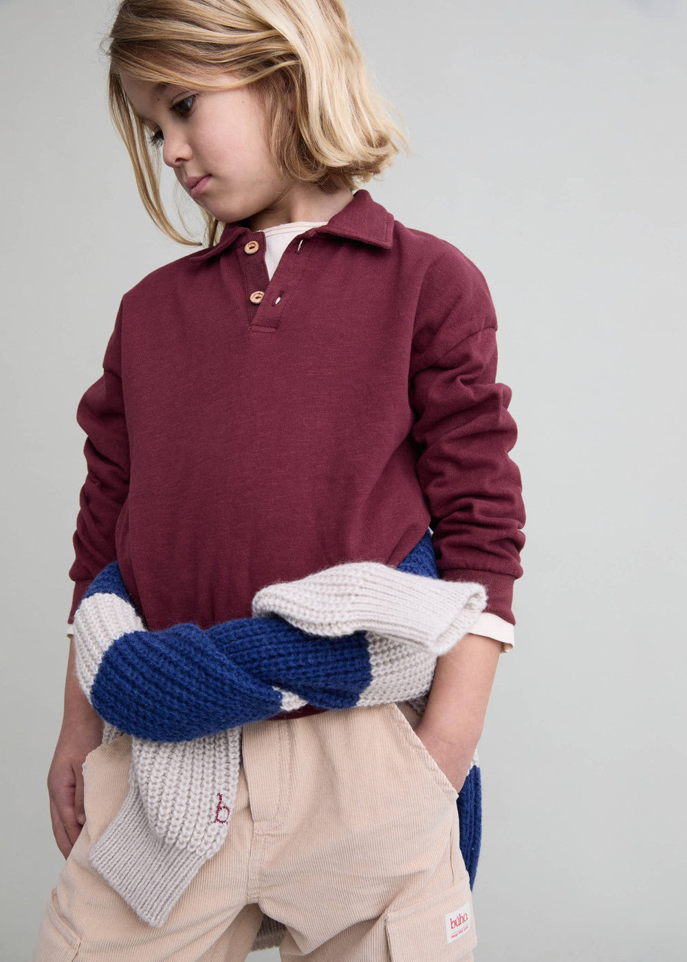 A child wearing a long-sleeve polo shirt in burgundy, paired with beige shorts and holding a blue and white knit blanket, against a plain light gray background.