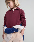 A child wearing a long-sleeve polo shirt in burgundy, paired with beige shorts and holding a blue and white knit blanket, against a plain light gray background.