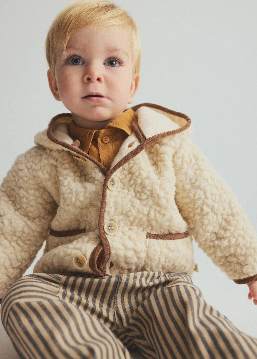 A baby wearing a cream sherpa fleece hooded jacket with brown trim, a mustard yellow shirt, and brown and beige striped pants, set against a plain light gray background.