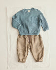A baby outfit set consisting of a light blue cable-knit long-sleeve top and beige-and-brown vertically striped pants with patch pockets, laid flat on a white sheet of paper against a light beige background.
