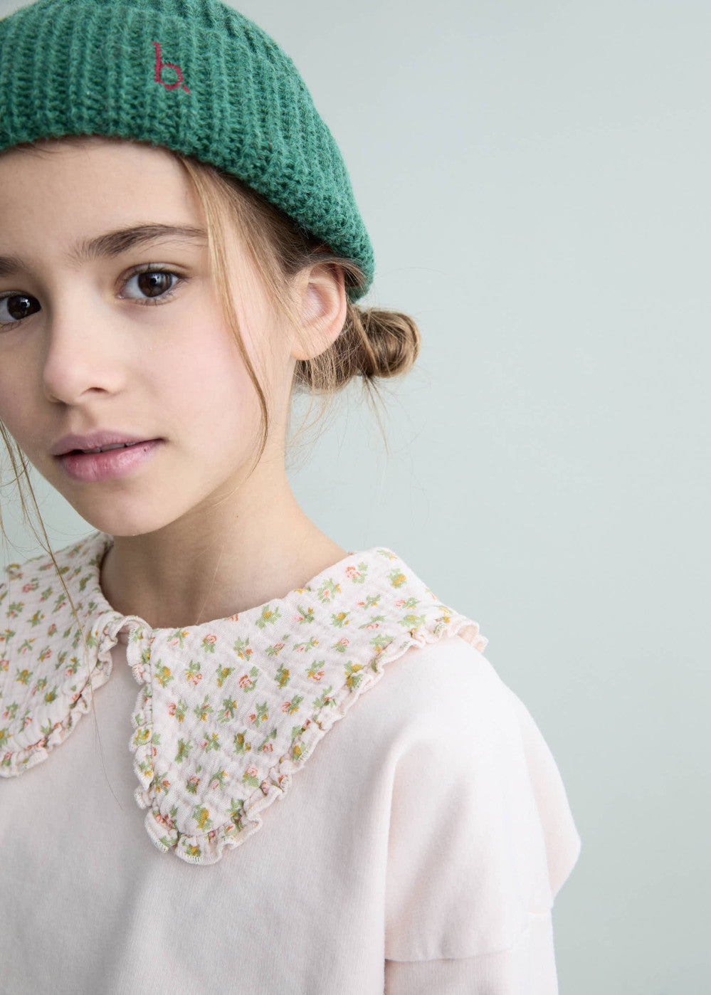 Young girl wearing a green knitted hat and a light pink blouse with a floral collar against a light gray background