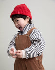 A smiling young child wearing a bright red knit beanie with a small white embroidered letter 'b', a black-and-white striped long-sleeve top, and brown corduroy overalls, posed against a plain light gray background.