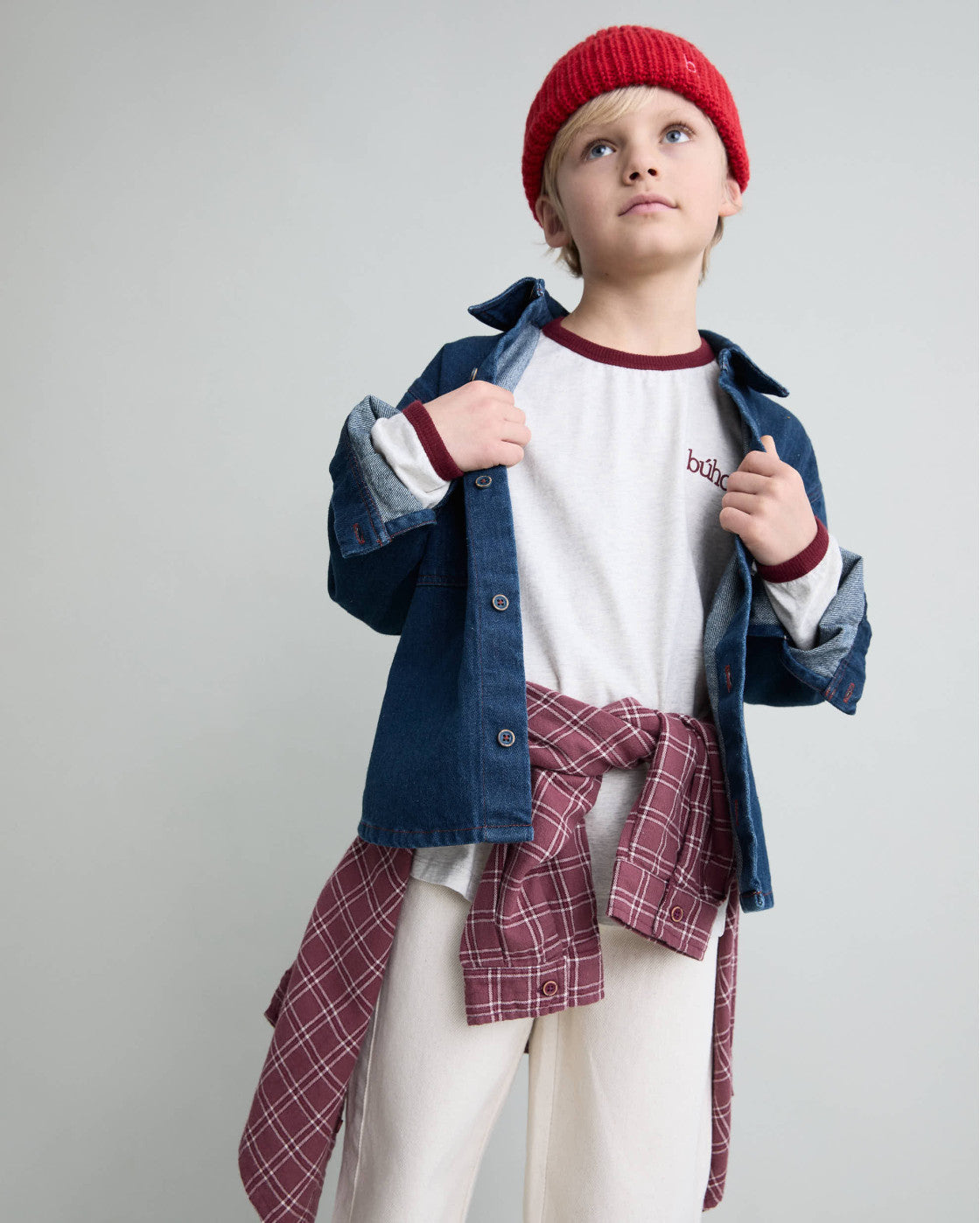 A child model wearing a red knit beanie, a white t-shirt with burgundy trim, a blue denim shirt-jacket, and a burgundy plaid shirt tied around the waist, set against a plain light gray studio background.