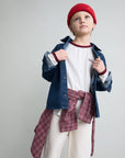 A child model wearing a red knit beanie, a white t-shirt with burgundy trim, a blue denim shirt-jacket, and a burgundy plaid shirt tied around the waist, set against a plain light gray studio background.