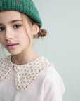 Young girl wearing a green knitted hat and a light pink blouse with a floral collar against a light gray background