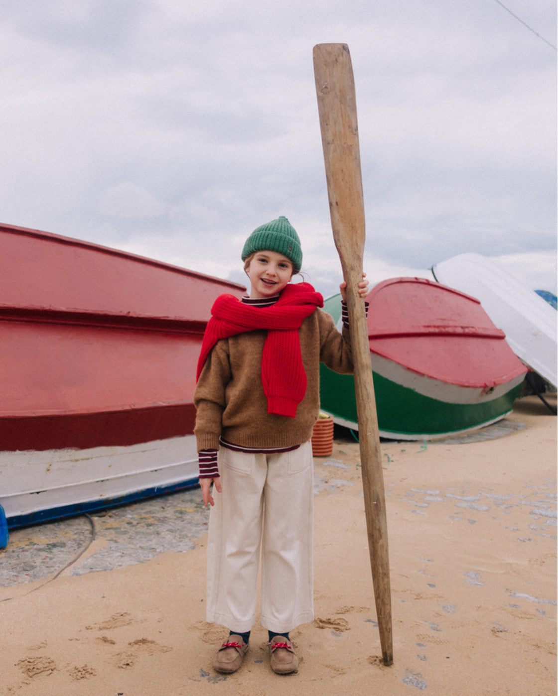 A child stands on a sandy beach holding a large wooden paddle, wearing a green knit beanie, a red scarf, a brown sweater, and cream wide-leg trousers, with red and green boats in the background under an overcast sky.