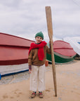 A child stands on a sandy beach holding a large wooden paddle, wearing a green knit beanie, a red scarf, a brown sweater, and cream wide-leg trousers, with red and green boats in the background under an overcast sky.