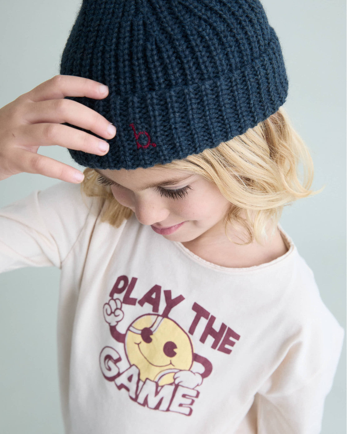 A child wearing a dark navy blue ribbed knit beanie with a small embroidered red logo and a cream graphic t-shirt, photographed against a light gray background.