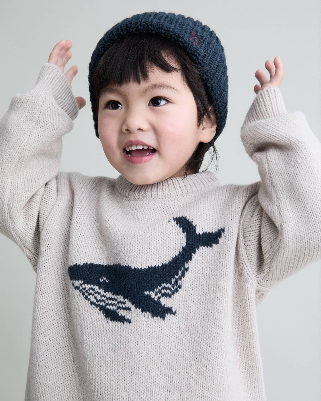 Child wearing a sweater with a whale design and a knit hat on a plain background