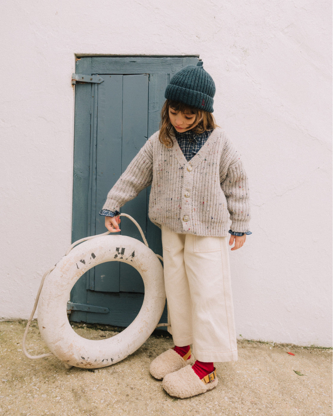 A young child stands outdoors in front of a white wall and blue door, wearing a beige knit cardigan, cream pants, a dark green beanie, and slippers, while holding a large white life preserver.