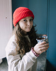 A young girl with long brown hair wearing a bright red knit beanie and a light gray sweatshirt, holding a small blue and white patterned cup in front of a dark blue paneled door.