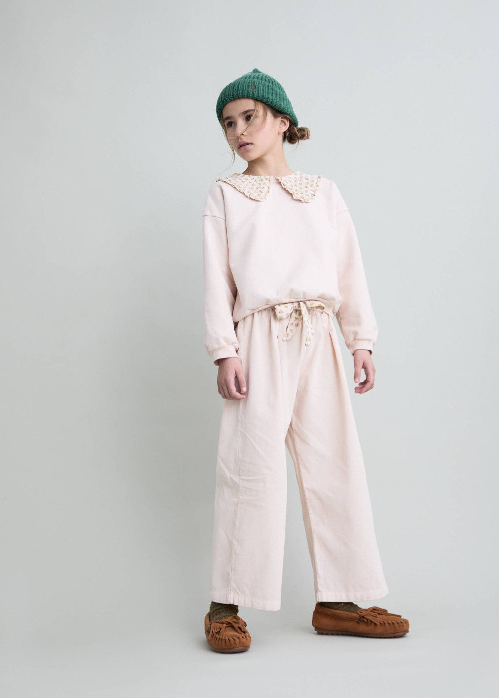 A child model wearing a light pink long-sleeve top with a large decorative collar, matching wide-leg trousers, a green knit beanie, and brown suede mule shoes, standing against a light gray studio background.