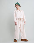 A child model wearing a light pink long-sleeve top with a large decorative collar, matching wide-leg trousers, a green knit beanie, and brown suede mule shoes, standing against a light gray studio background.