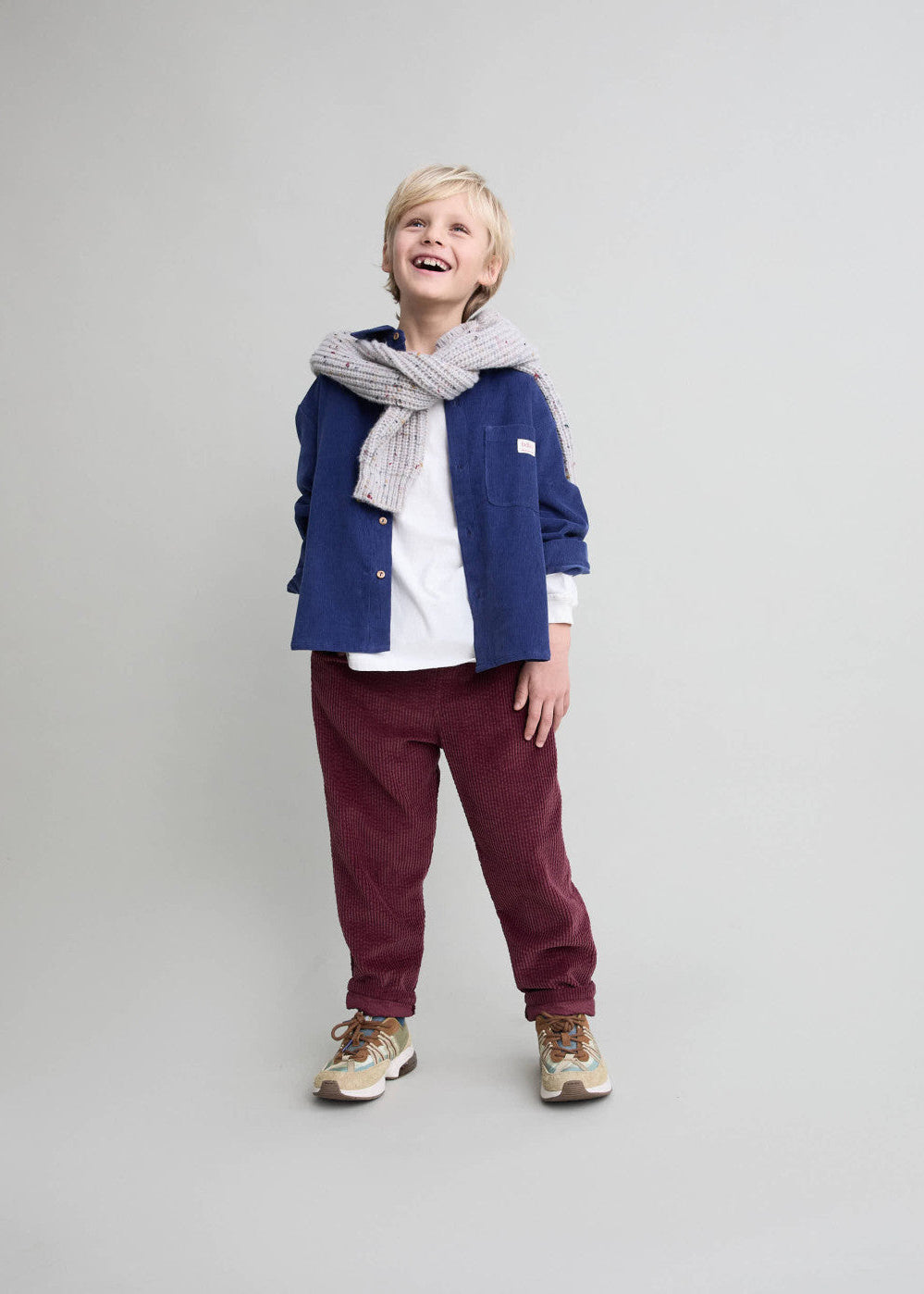 Child wearing a blue jacket, white shirt, maroon pants, and gray scarf on a light gray background
