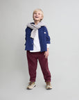 Child wearing a blue jacket, white shirt, maroon pants, and gray scarf on a light gray background