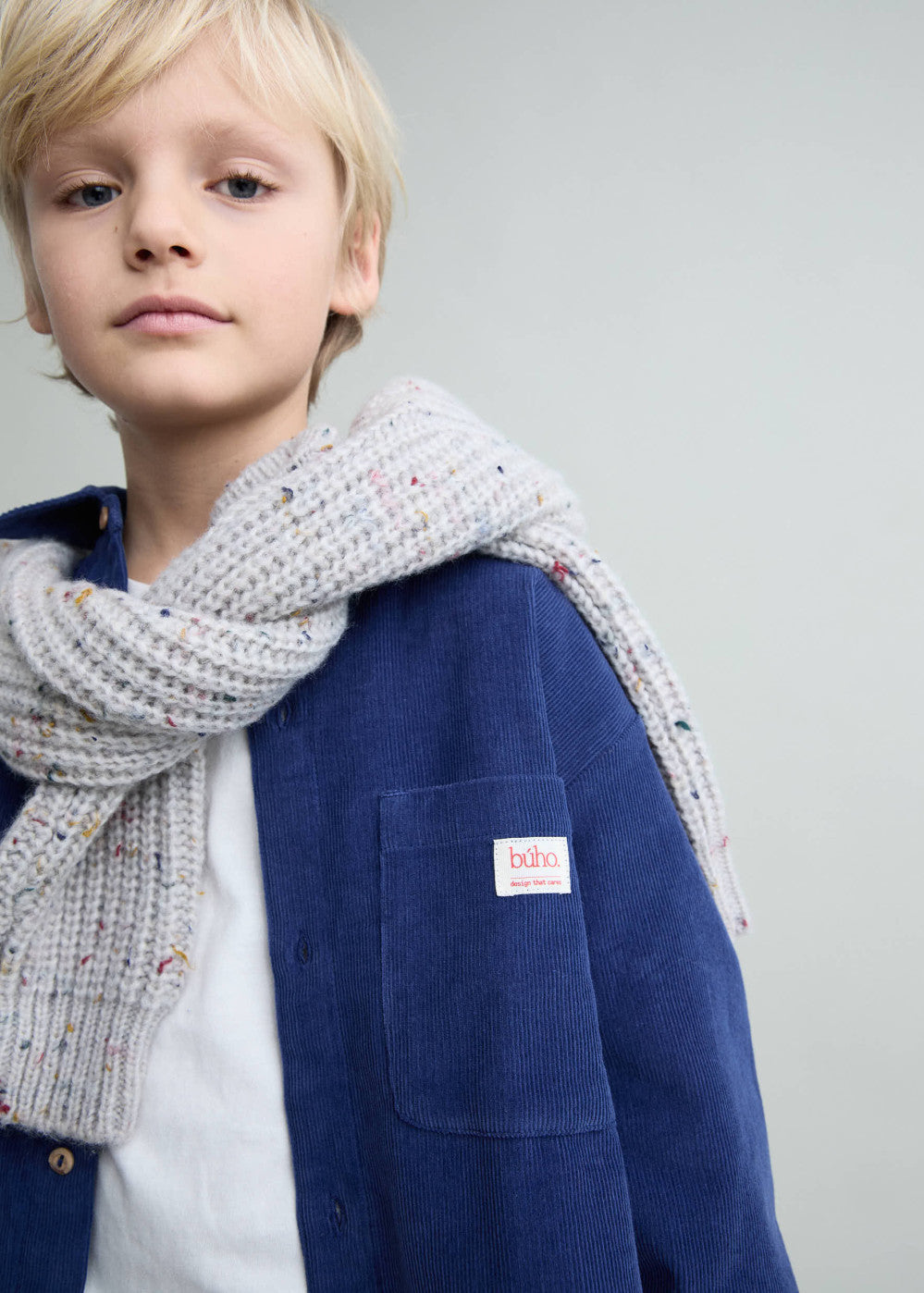 A young blonde boy wearing a navy blue corduroy jacket, a white t-shirt, and a chunky light gray knit scarf with tiny multicolor flecks. The jacket features a small white patch on the chest pocket with the brand name 'baba berlin'.
