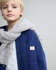 A young blonde boy wearing a navy blue corduroy jacket, a white t-shirt, and a chunky light gray knit scarf with tiny multicolor flecks. The jacket features a small white patch on the chest pocket with the brand name 'baba berlin'.