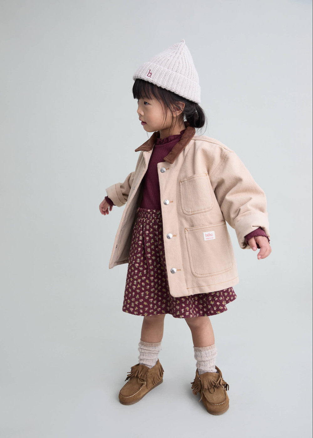 Child wearing a beige coat, maroon dress, white hat, and brown shoes on a light gray background