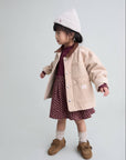 Child wearing a beige coat, maroon dress, white hat, and brown shoes on a light gray background