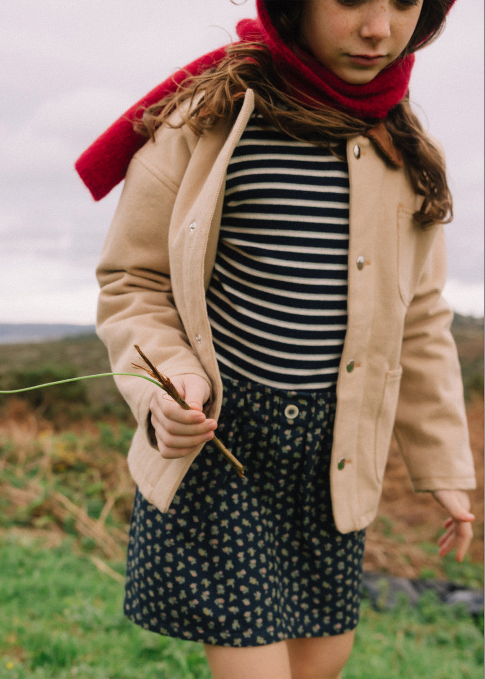 A child wearing a beige jacket over a navy and white striped top and a navy floral skirt, with a bright red scarf, standing outdoors in a grassy field under an overcast sky.