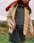 A child wearing a beige jacket over a navy and white striped top and a navy floral skirt, with a bright red scarf, standing outdoors in a grassy field under an overcast sky.