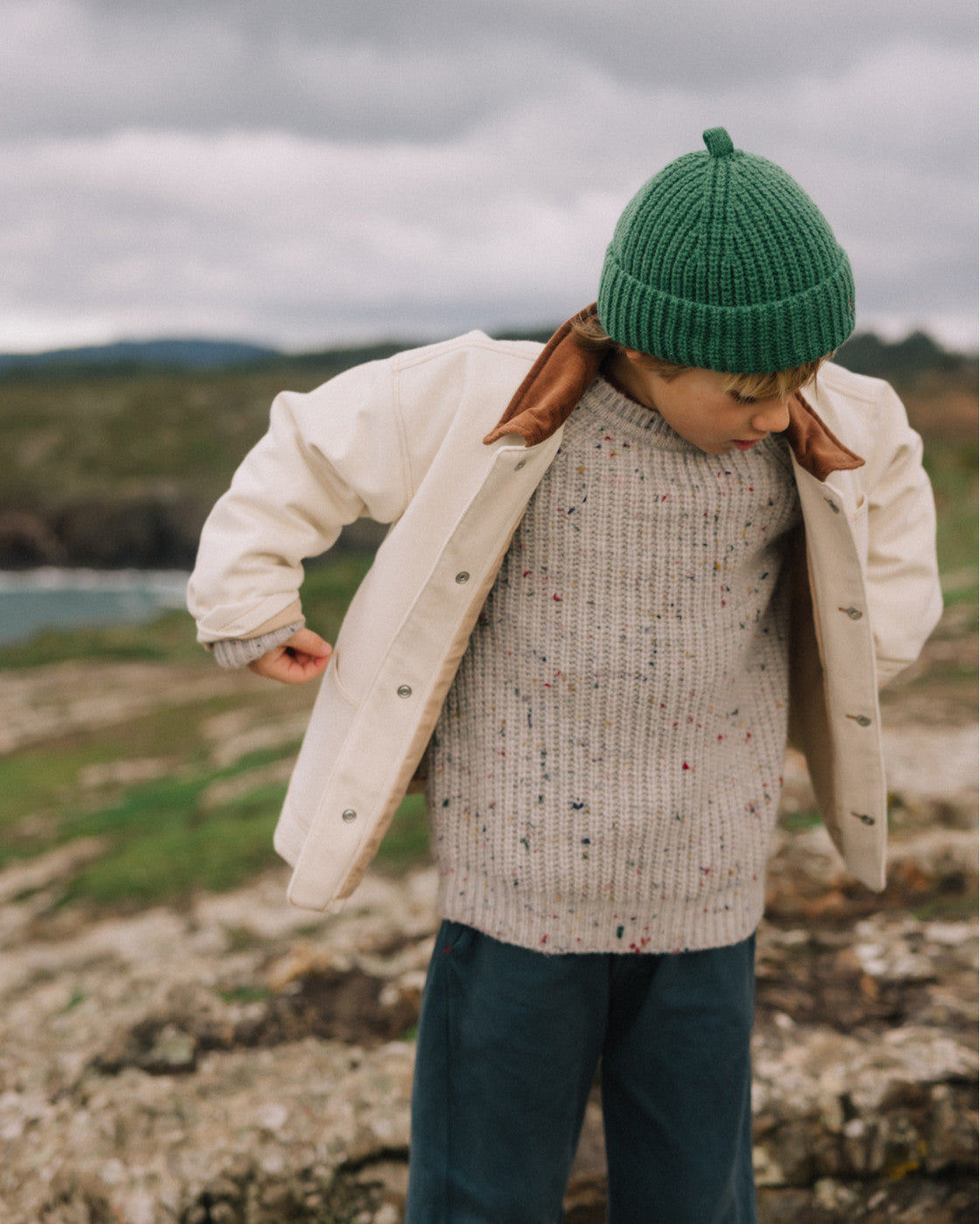 A child wearing a cream jacket with a brown shearling-style collar, a speckled beige knit sweater, a green beanie, and dark teal pants, standing outdoors on a rocky shoreline under an overcast sky.