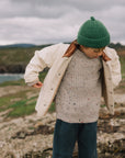 A child wearing a cream jacket with a brown shearling-style collar, a speckled beige knit sweater, a green beanie, and dark teal pants, standing outdoors on a rocky shoreline under an overcast sky.
