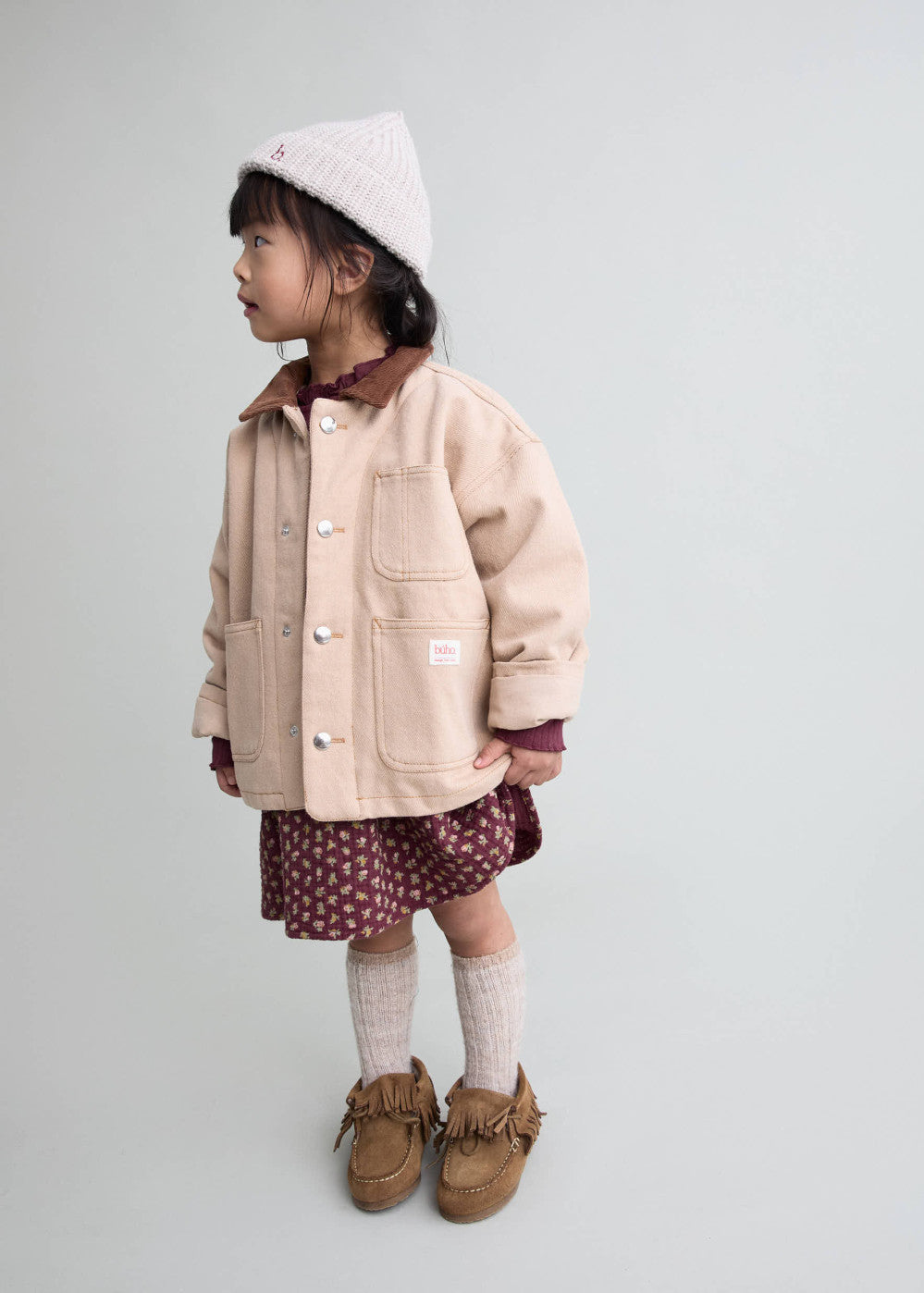 A young child stands in profile wearing a light beige jacket, a burgundy patterned skirt, cream socks, brown shoes, and a cream knit beanie against a plain light gray studio background.