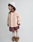 A young child stands in profile wearing a light beige jacket, a burgundy patterned skirt, cream socks, brown shoes, and a cream knit beanie against a plain light gray studio background.