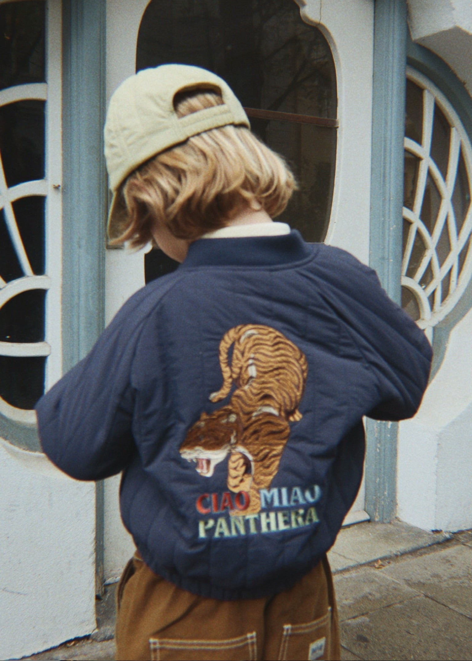 Person wearing a navy blue jacket with a tiger design and 'CIAO MIAO PANTEONA' text, standing in front of a decorative door.