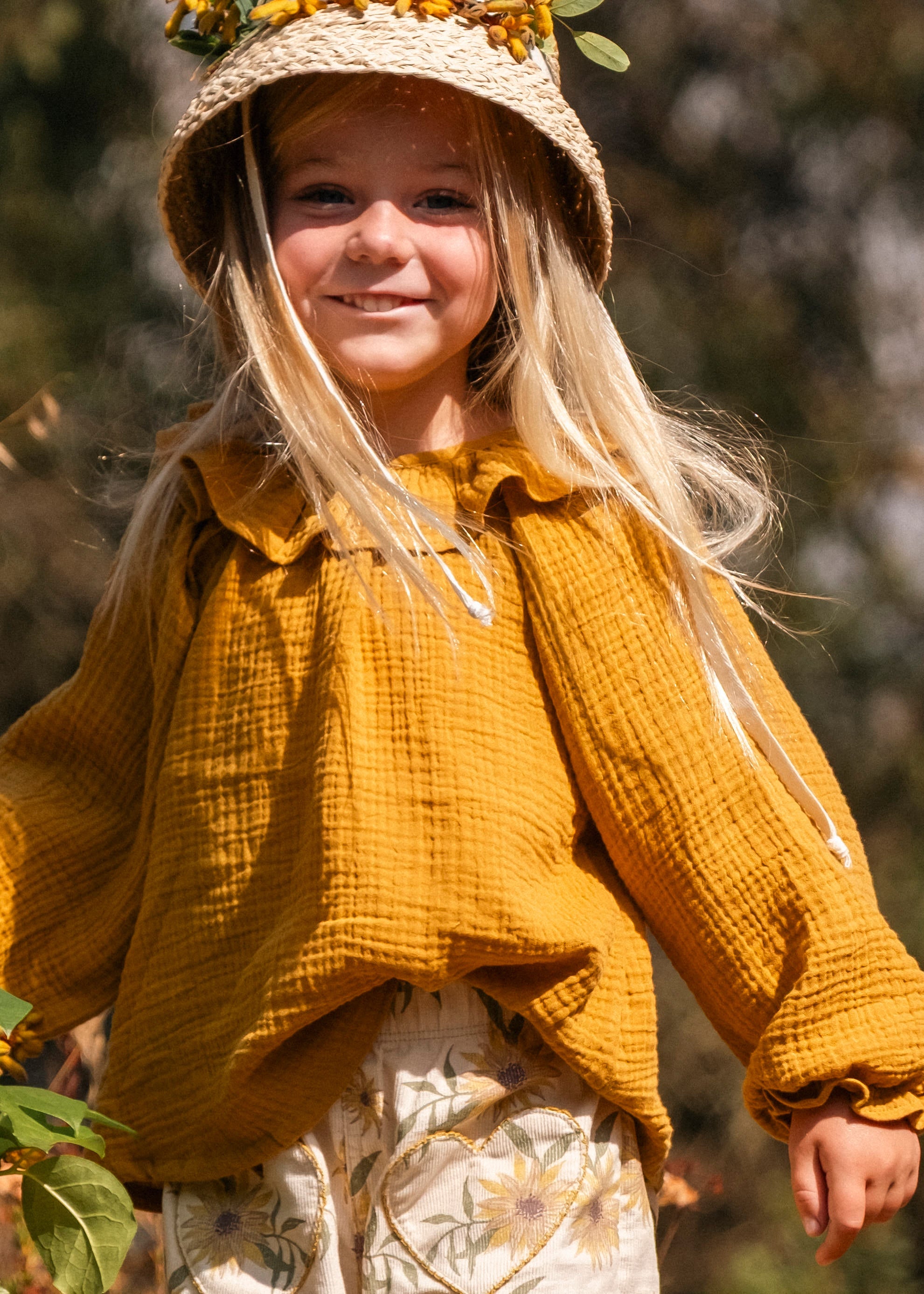 Child in a yellow outfit with a hat, standing outdoors among plants.